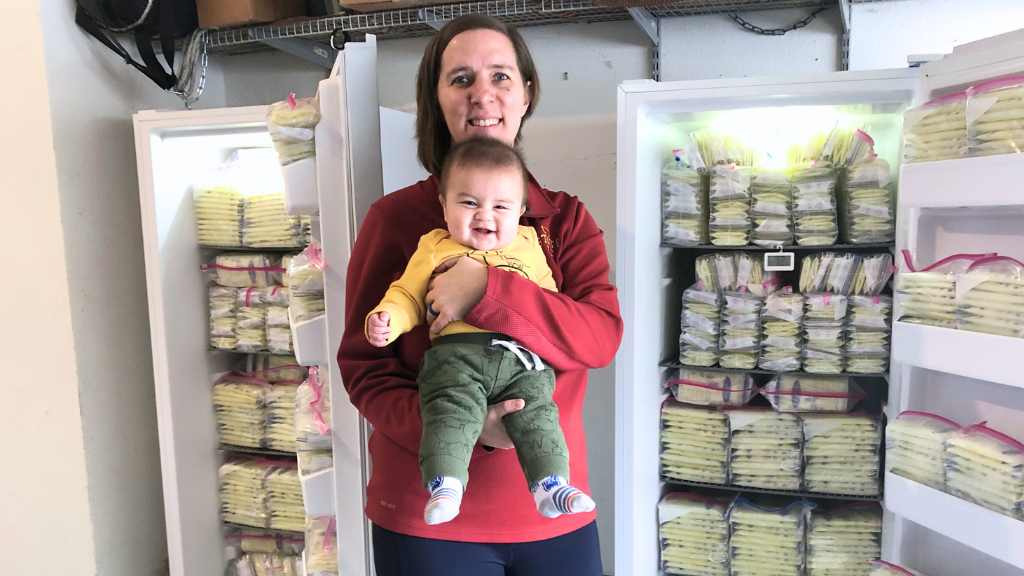 surrogate holding surrogacy baby in front of two freezers full of pumped breastmilk