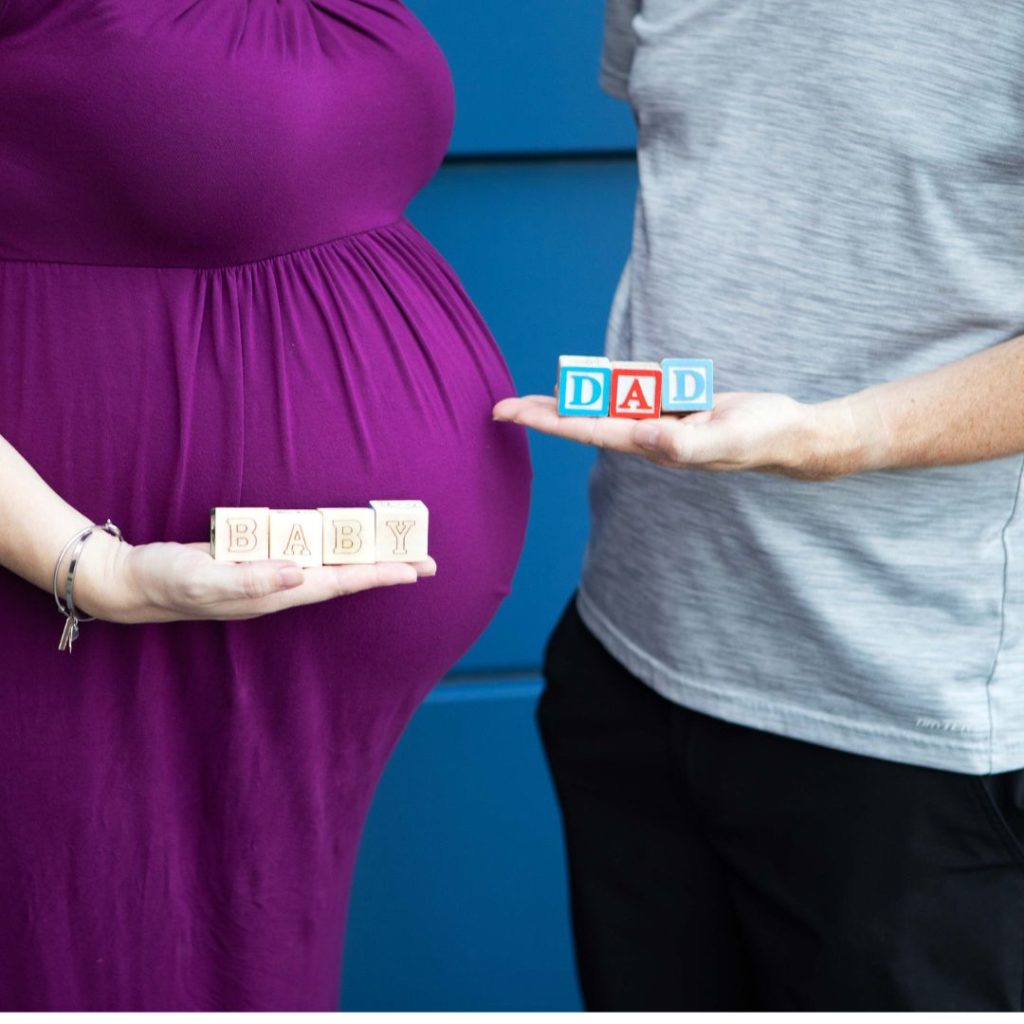 surrogacy maternity shoot with surrogate holding wooden blocks that spell "baby" and intended single father holds wooden blocks that spell "dad"