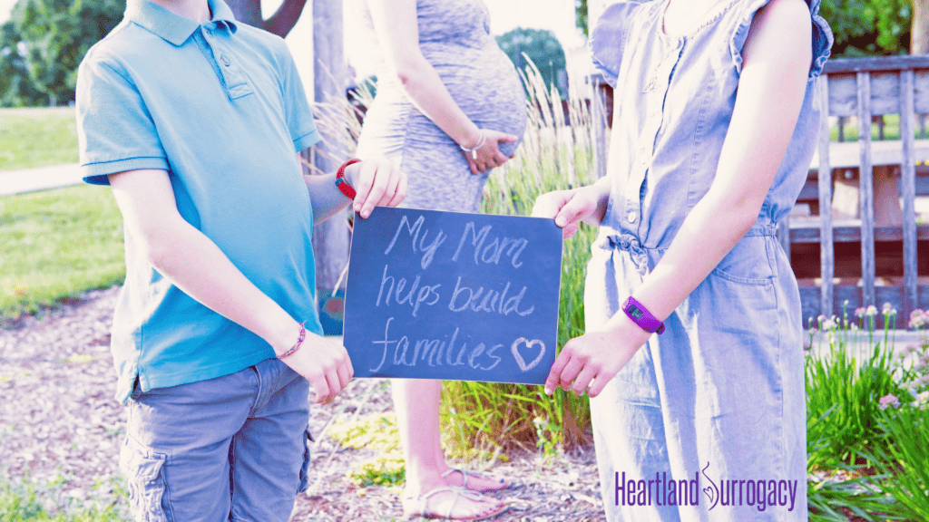 Iowa surrogate mother with family and sign reading “My mom helps build families”.]